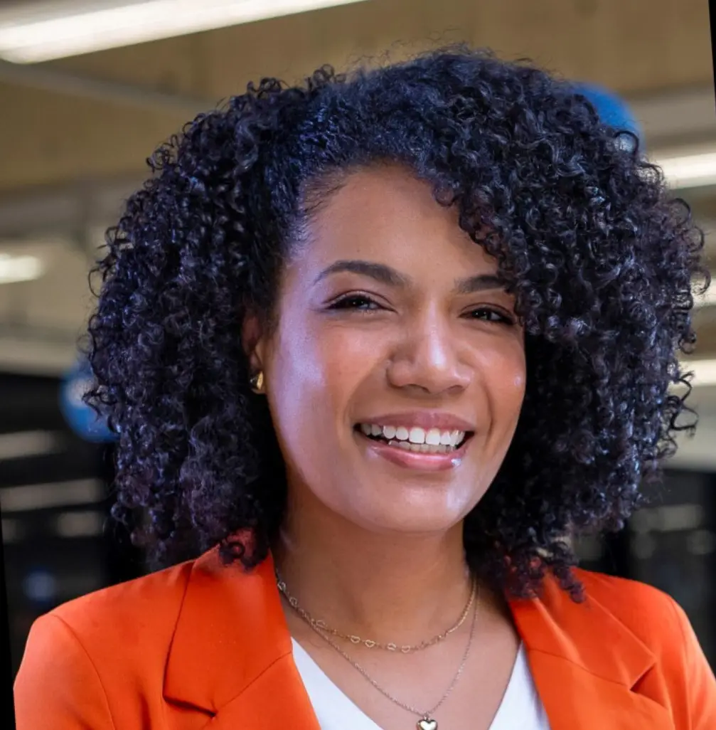 Foto de Camila Ribeiro uma mulher usando um blazer laranja e camisa branca, com cabelos cacheados na altura dos ombros.